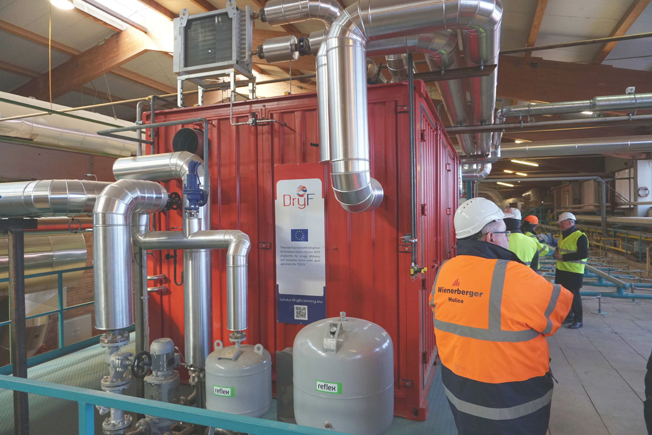 A large industrial red container unit with complex piping and machinery is viewed from the side, with several people in hard hats and high-visibility vests looking at it.