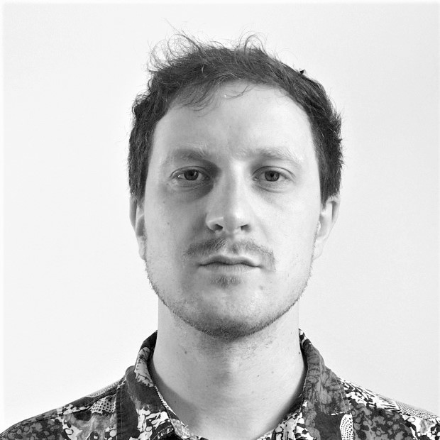 A person with short hair wearing a patterned shirt stands against a plain white background, with an obscured face.
