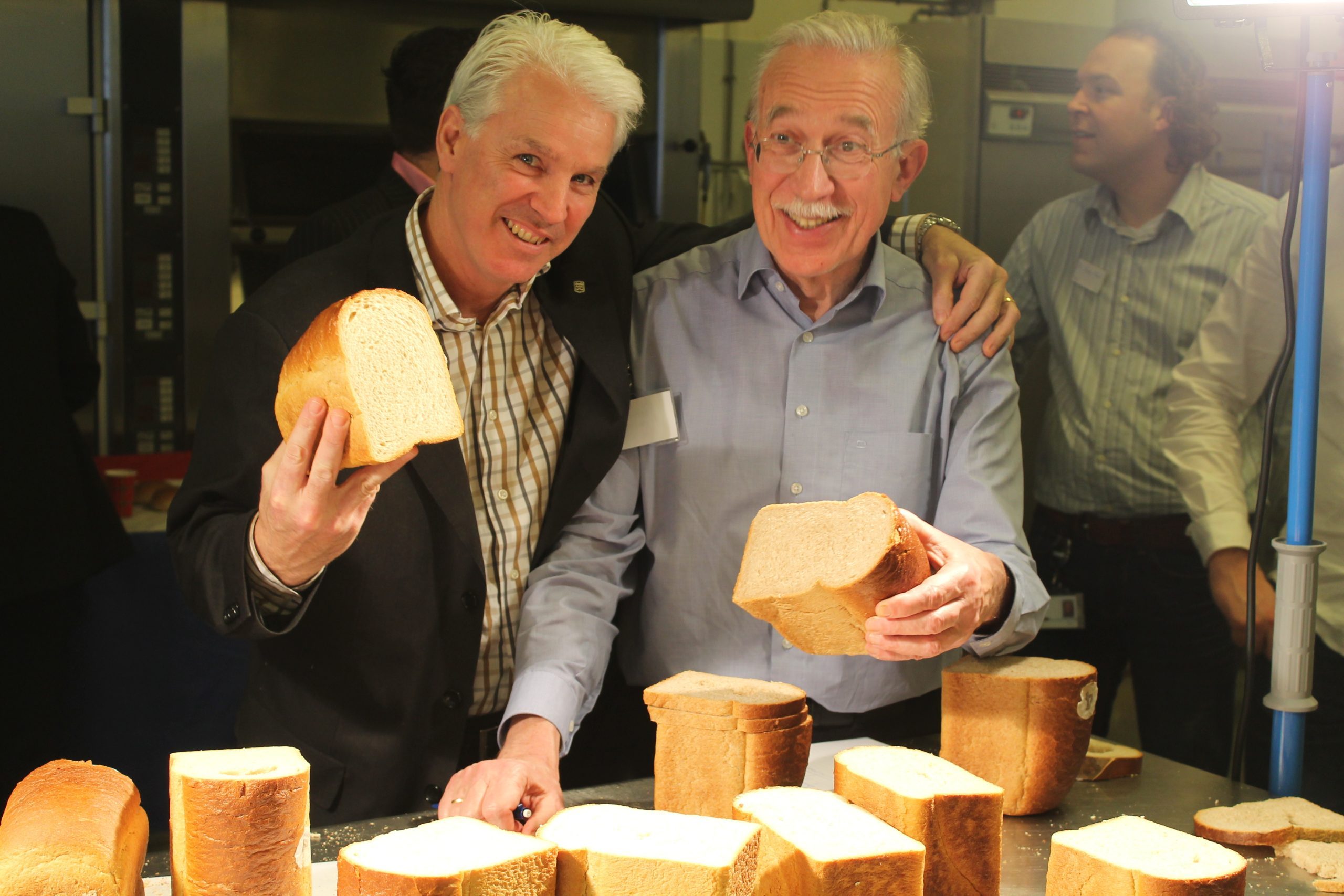 Two individuals hold slices of freshly baked bread, surrounded by loaves on a table in a bustling kitchen environment.