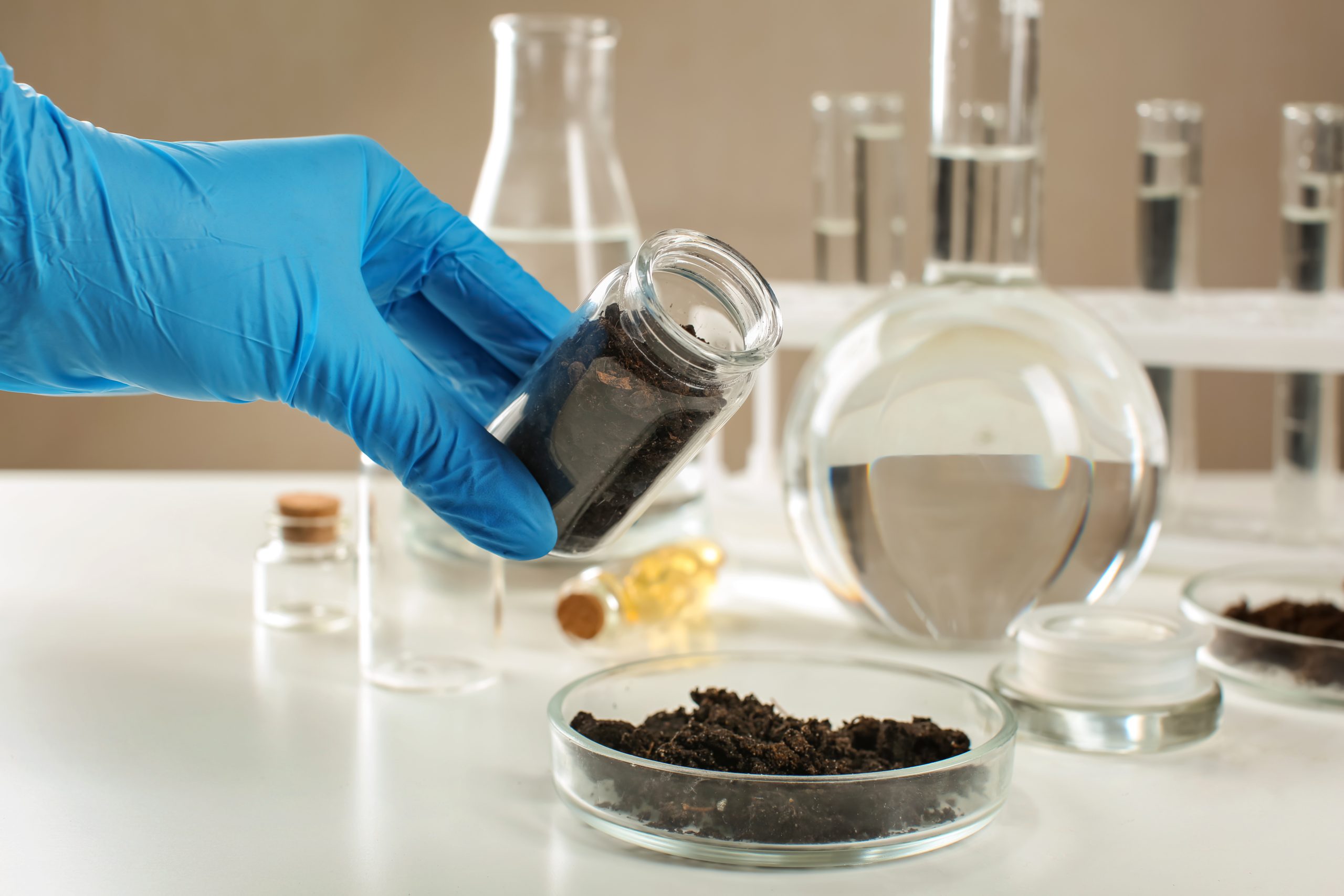 A gloved hand holds a jar of soil above a petri dish filled with soil, surrounded by various lab glassware.