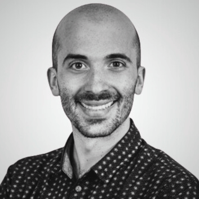 A close-up portrait of an individual wearing a patterned shirt, captured in black and white against a neutral background.