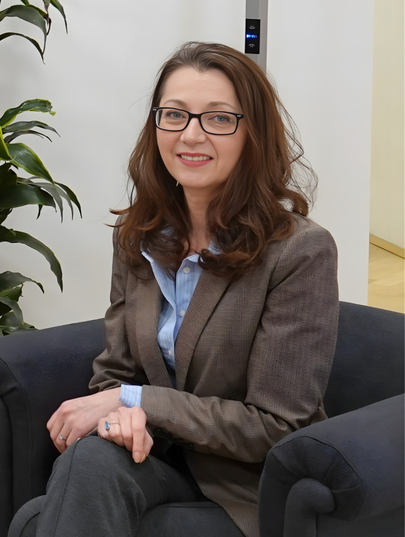 Roxana Weiss Anton, CEO of RTDS Group Professional headshot of a woman with long brown hair and glasses, wearing a brown blazer over a light blue button-down shirt. She is sitting in a dark gray armchair and smiling in an office setting.